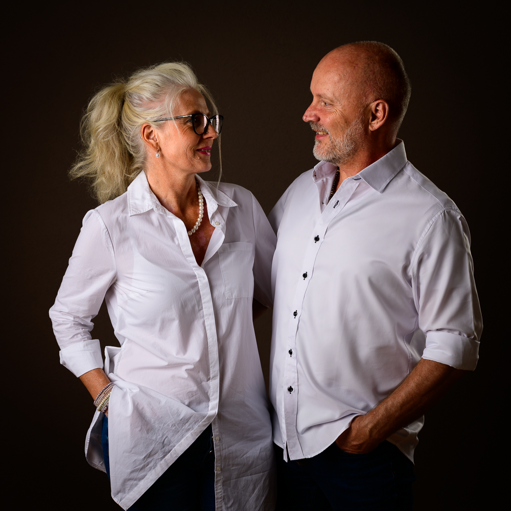 Über uns - authentisch und kreativ Nicole und Felix stehen nebeneinander in einem Raum mit warmer Beleuchtung. Sie lächeln einander an und vermitteln die Authentizität und laden ein, ein einzigartiges Erlebnis mit ihnen durch MindArt@KIWI einzugehen.